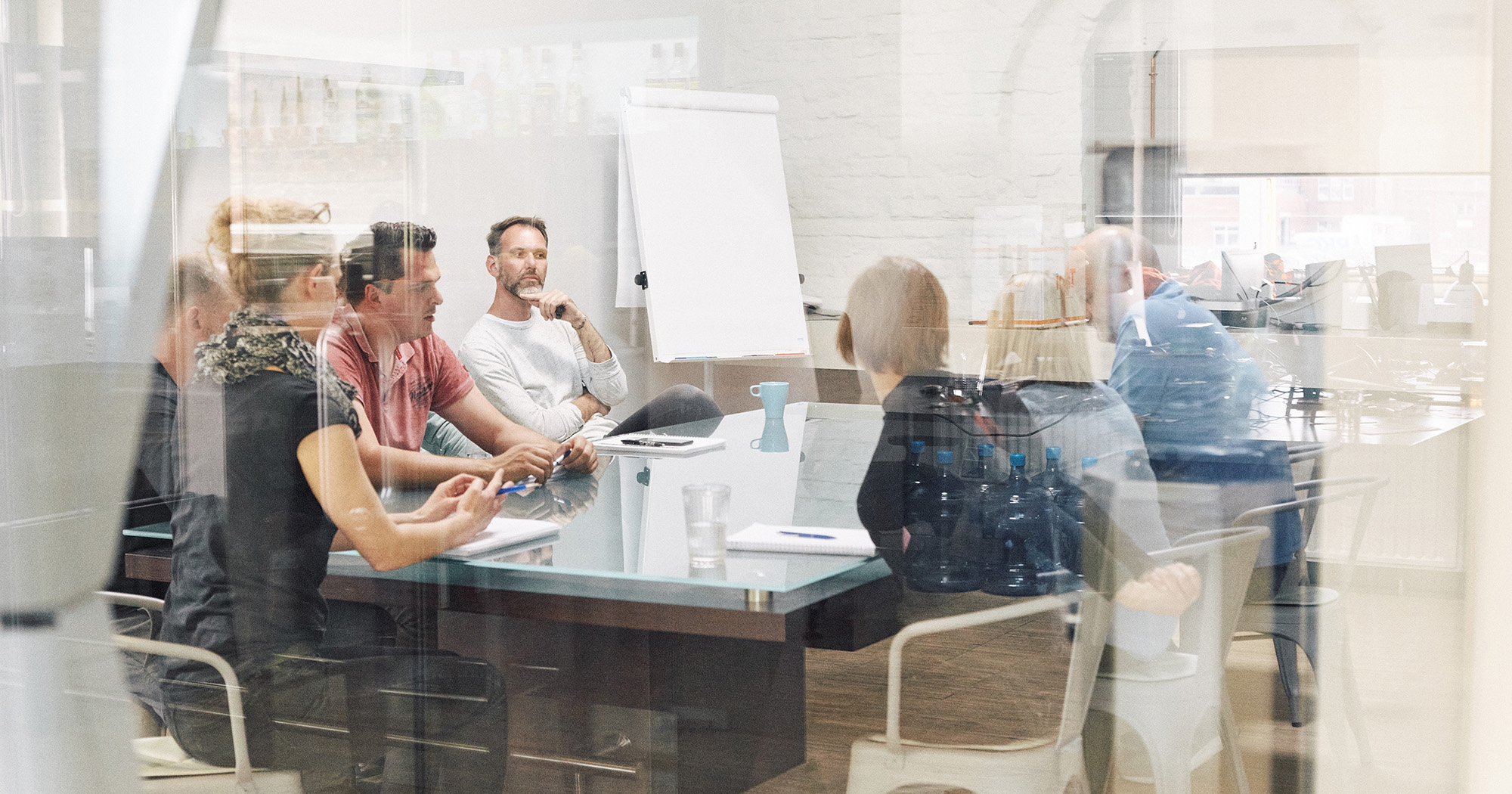 Man sieht mehrere Mitarbeiter von FederhenSchneider unterschiedlichen Geschlechts und Alters in einem Konferenzraum durch die Scheibe fotografiert. Sie sehen nachdenklich aus.