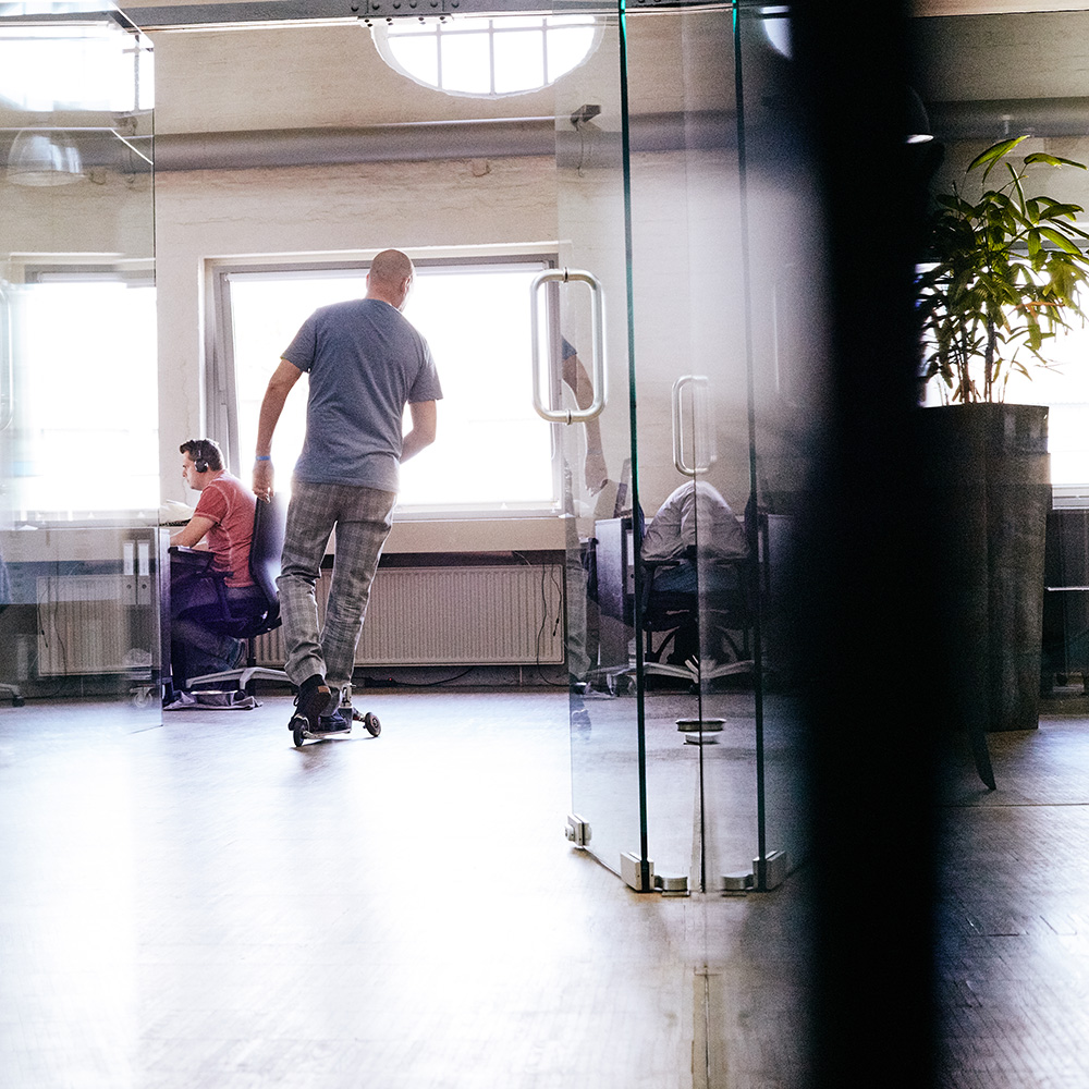Das Bild zeigt einen der FederhenSchneider Geschäftsführer von hinten, wie er auf einem Cityroller durch den Gang der Agentur fährt.