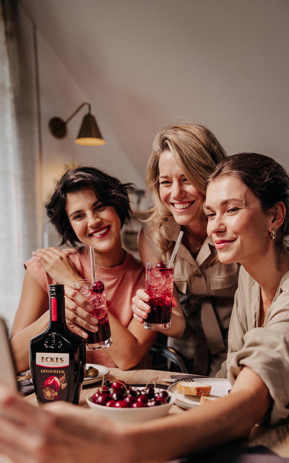 Drei Frauen machen ein Selfie und halten Cocktails aus ECKES Edelkirsch in der Hand. Eine Flasche ECKES Edelkirsch steht auf dem Tisch.