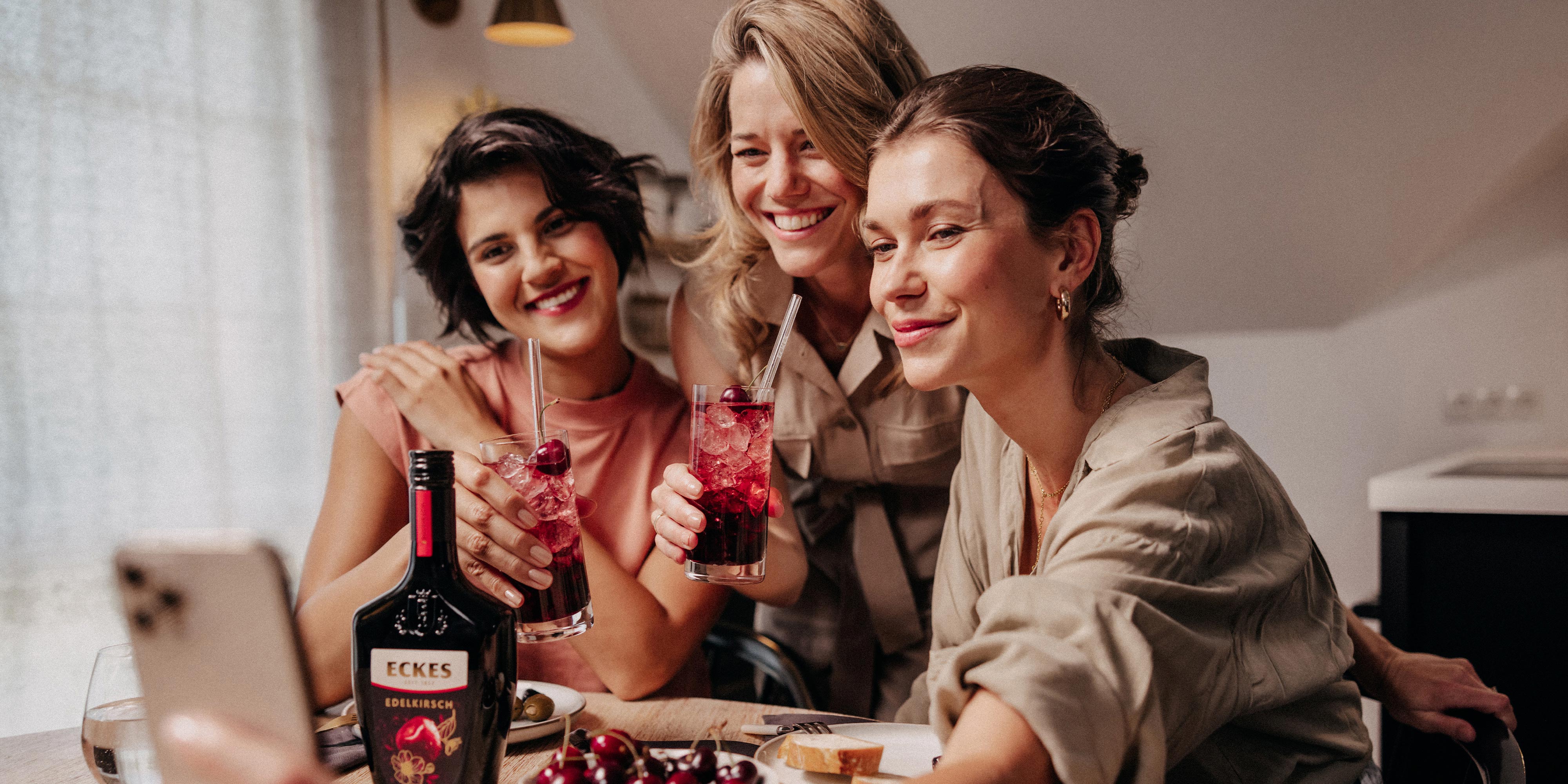 Drei Frauen machen ein Selfie und halten Cocktails aus ECKES Edelkirsch in der Hand. Eine Flasche ECKES Edelkirsch steht auf dem Tisch.